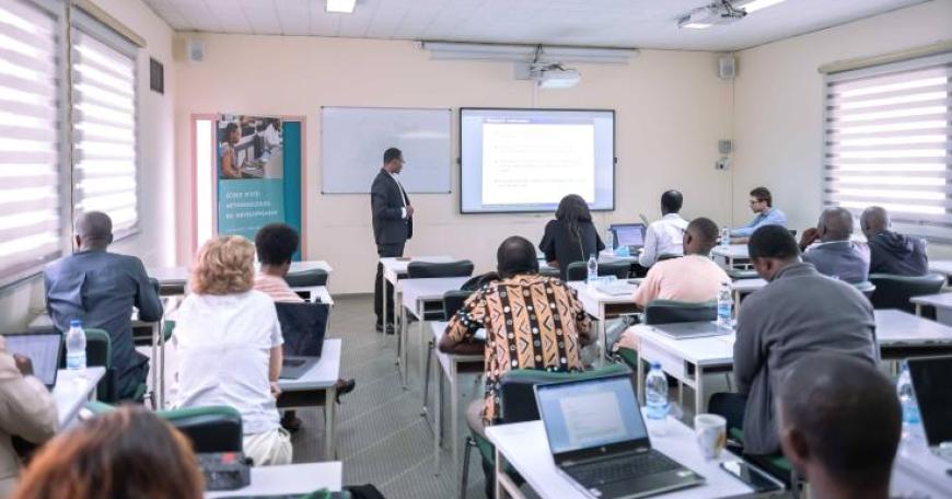 Candid photo of a classroom with over a dozen adults seated at desks with open laptops. An instructor stands at the front to explain the lesson displayed on the screen on the wall.